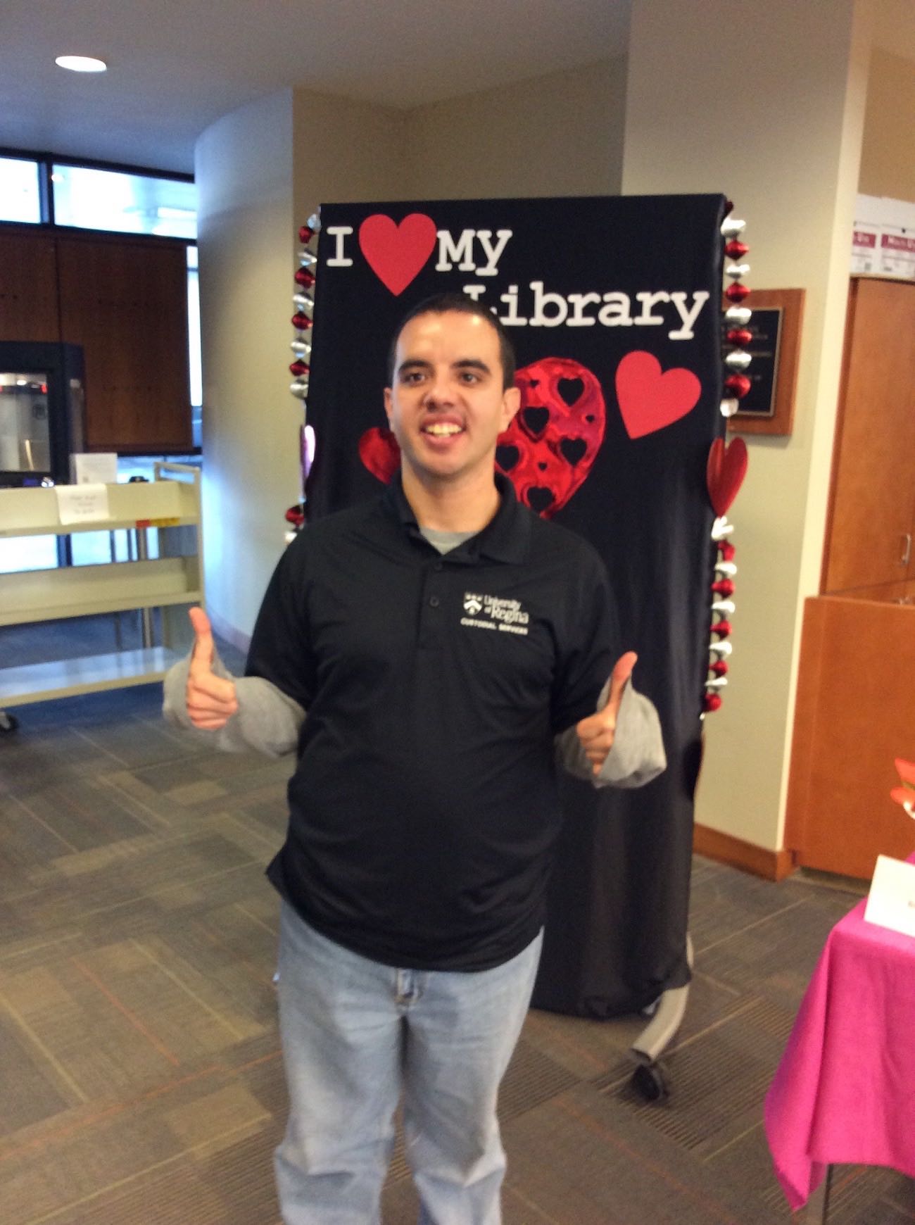 One of the library's Facilities Management staff stands in front of the I love my Library backdrop smiling and giving two thumbs up